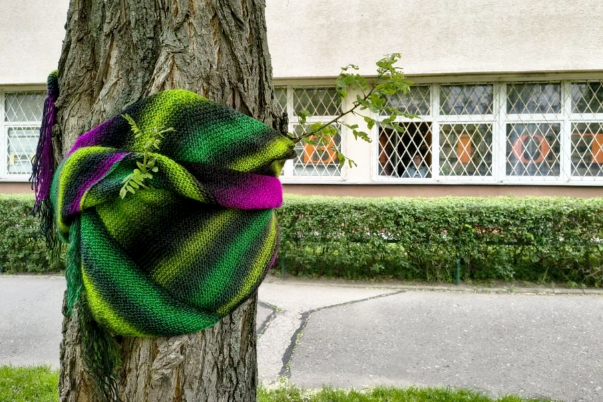 Drzewo owinięte kolorowym, wełnianym szalem w zielono-fioletowe paski, przymocowanym do pnia. W tle widoczny budynek z dużymi oknami i zielony żywopłot, a także fragment drogi.